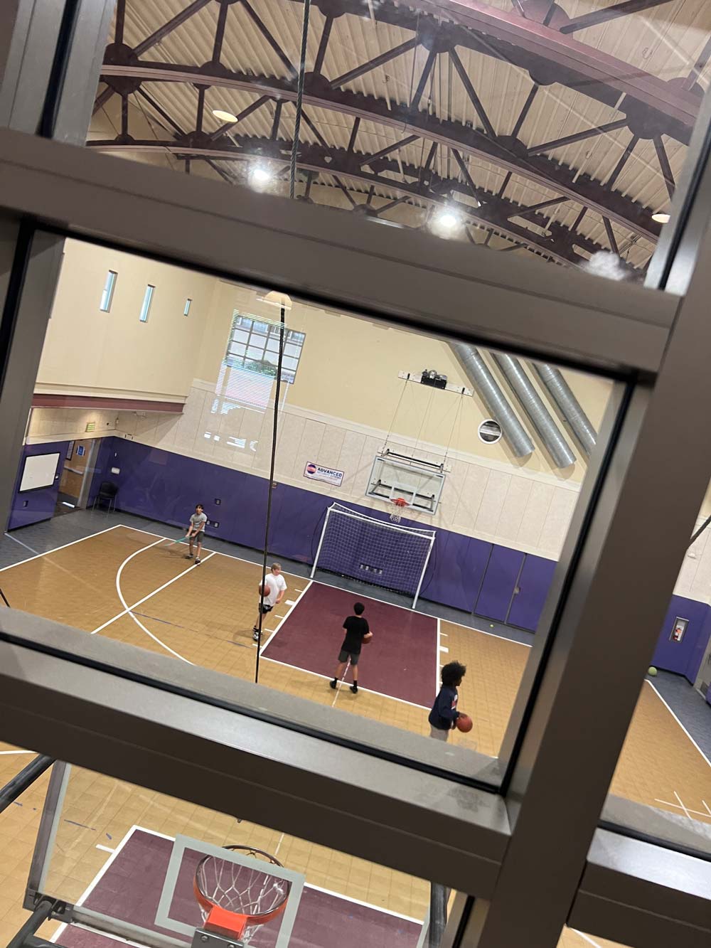 View through a window of an indoor basketball court with four people playing and a basketball hoop in the foreground.