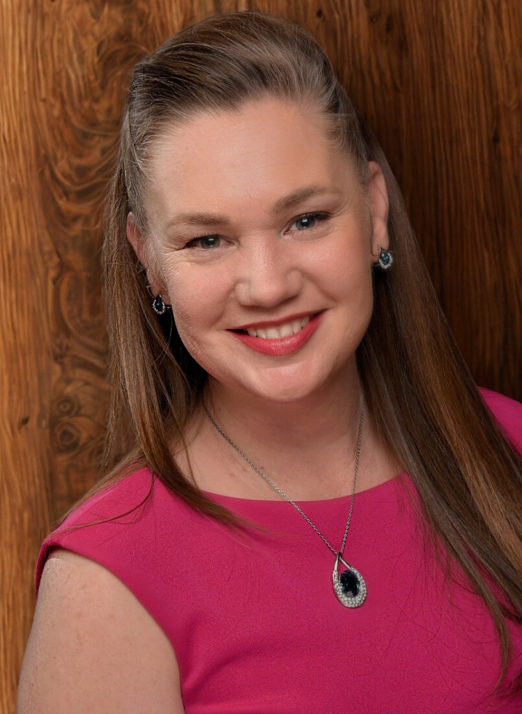 Woman with long brown hair wearing a pink sleeveless dress and a pendant necklace, smiling in front of a wooden background.