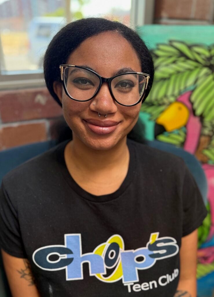 A person with glasses and a nose ring sits in a chair, wearing a black "chops Teen Club" T-shirt. A colorful tropical painting is visible in the background.