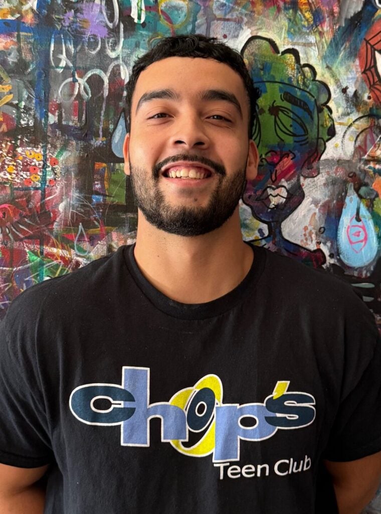 A man with a beard smiles in front of a colorful, abstract graffiti-style painting. He is wearing a black "chops Teen Club" t-shirt.
