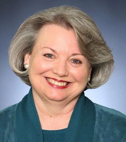Smiling person with short gray hair wearing a blue top, positioned against a neutral background.