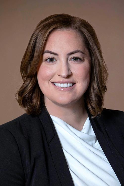 Person with shoulder-length brown hair, wearing a black blazer and white top, smiling against a plain brown background.