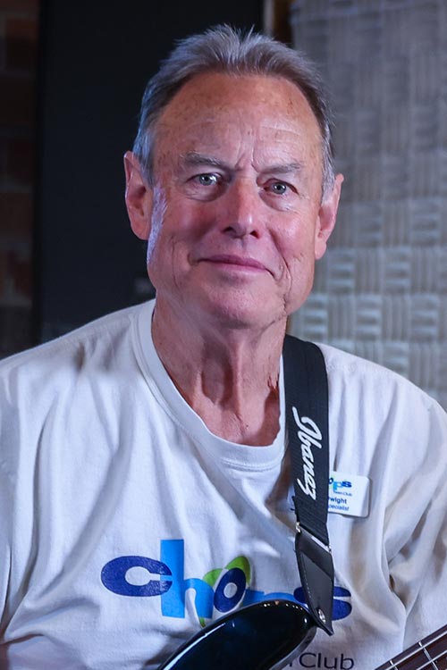 An older man with gray hair plays a black bass guitar, wearing a white t-shirt and a name badge, with a neutral expression.