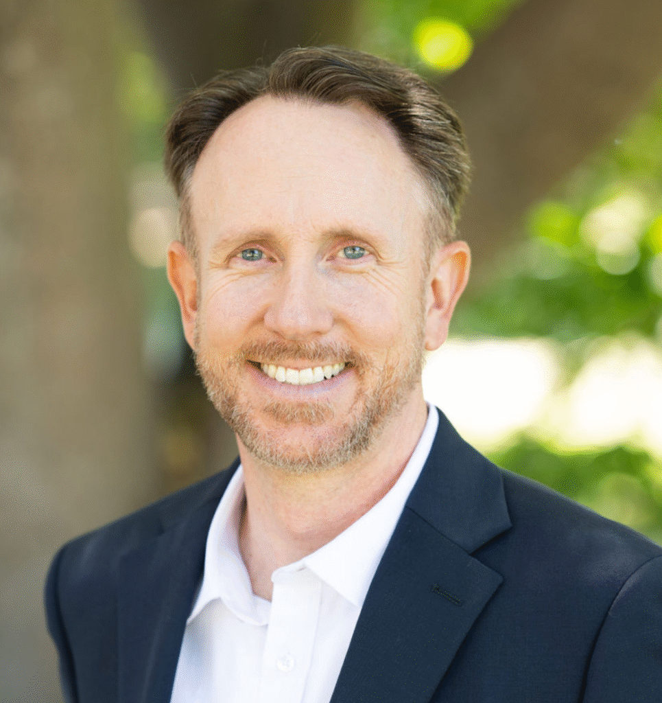 A man with short brown hair and a beard, wearing a dark blazer and white shirt, smiles outdoors with trees in the background.