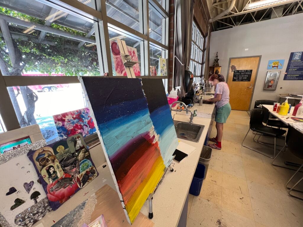 Painting on an easel with colorful stripes sits in an art classroom. A person is working near a sink on the right.