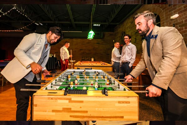 Two men playing foosball while others watch in a casual indoor setting.