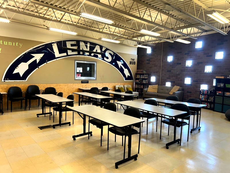 A classroom with tables and chairs arranged in rows, a large sign reading "LENAS" on the wall, bookshelves, a window, and exposed ceiling beams.