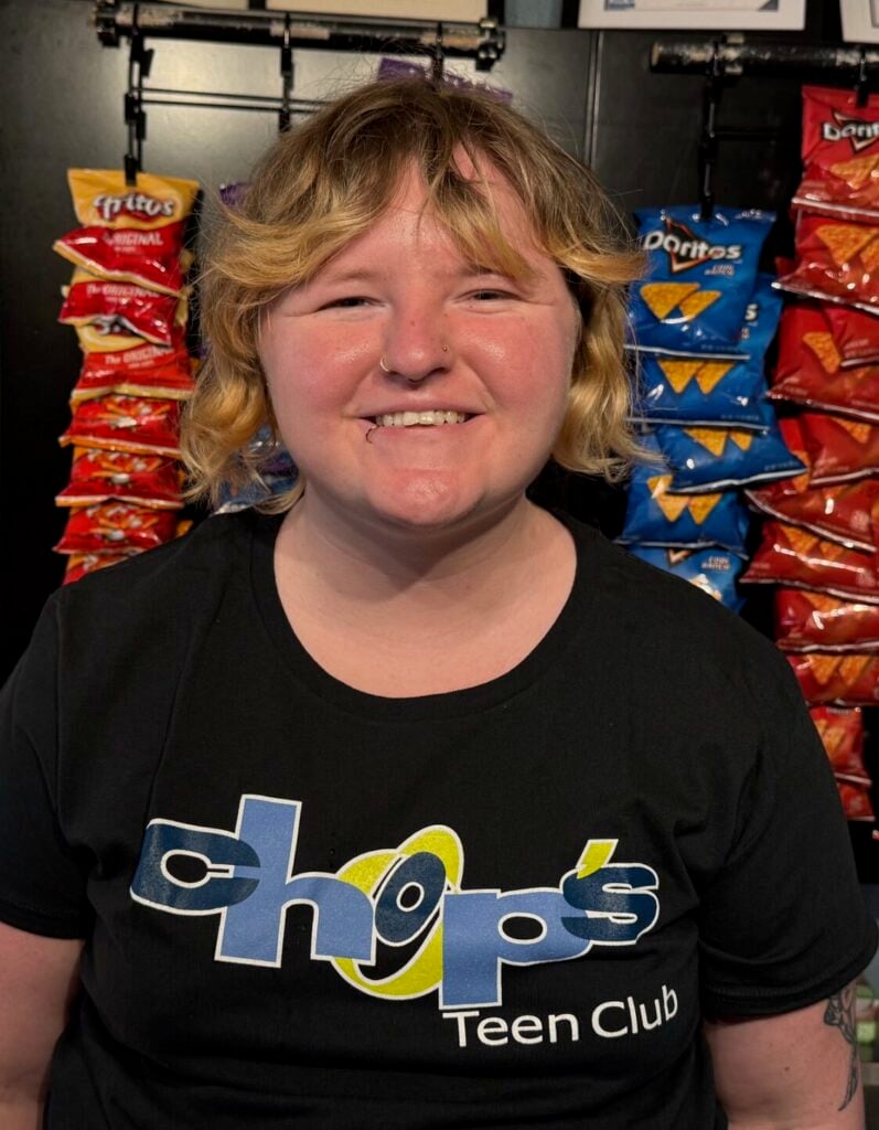 A person with short, wavy hair smiles while standing in front of a display of chips. They are wearing a black "Chops Teen Club" t-shirt. Framed certificates are on the wall above.