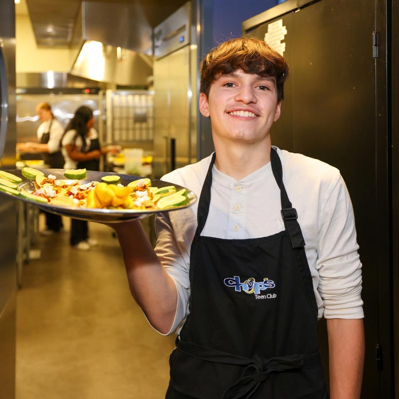 A young person in an apron holds a platter of food in a kitchen. Other people are working in the background.