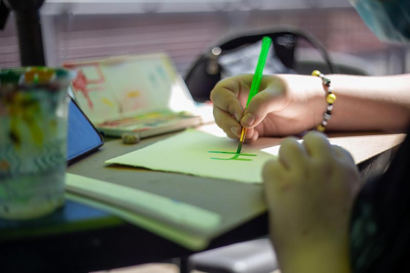 Hand drawing a green hashtag on paper with a green pen. Nearby are a paint palette, a cup with brushes, and a smartphone.