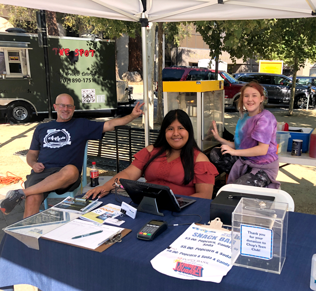 Three people sit at a snack bar booth outdoors with paperwork, a calculator, and a popcorn machine under a canopy at an event.