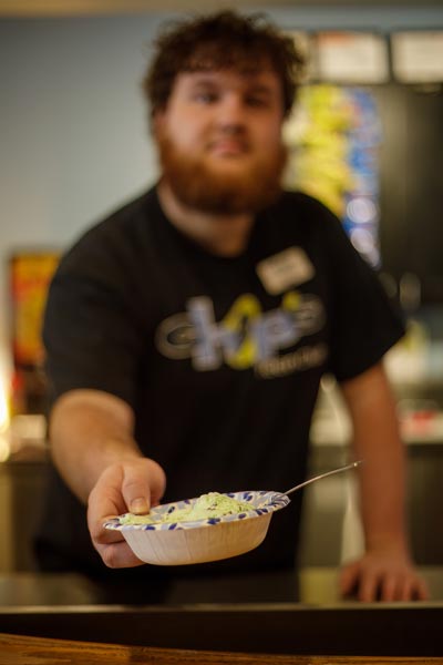 A person with a beard offers a bowl of ice cream with a spoon, standing behind a counter indoors.