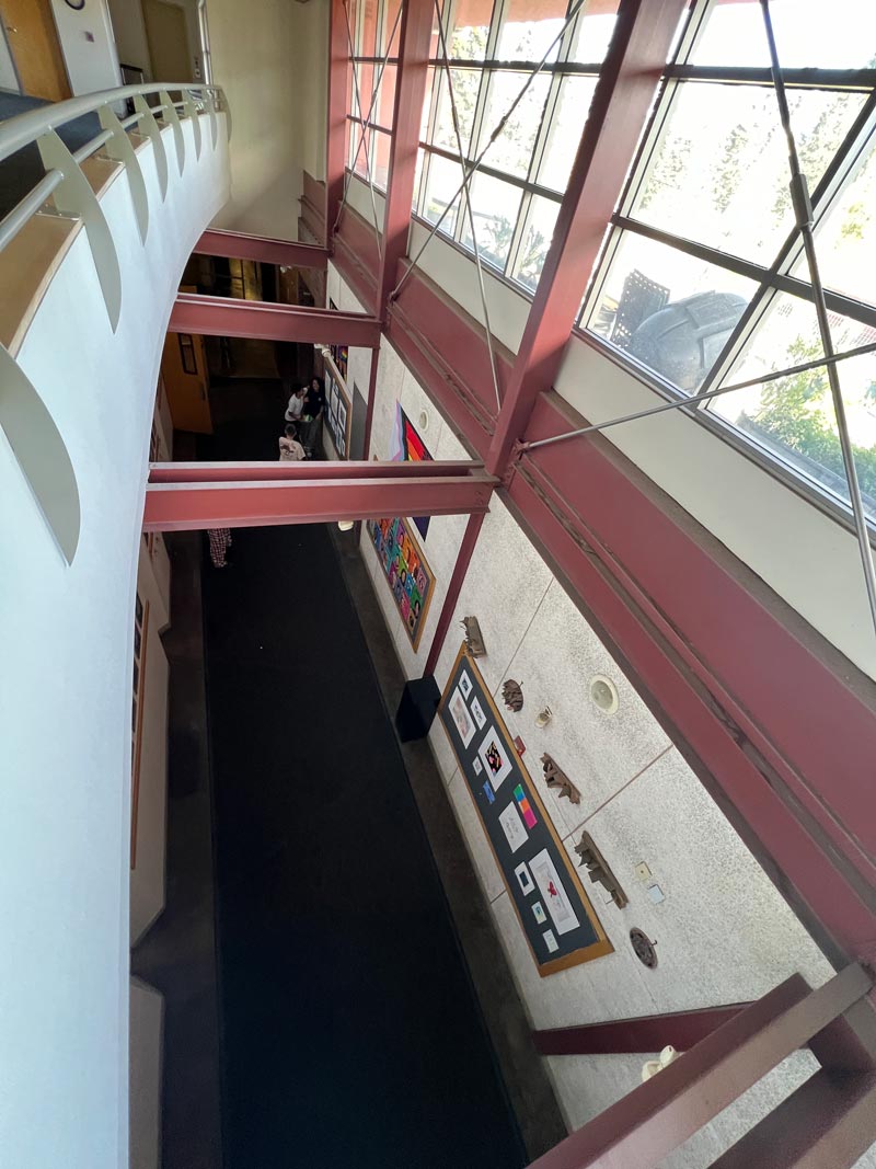 View from above of a hallway with a curved wall to the left and large windows on the right. Framed artwork is displayed on the walls below. Red beams cross overhead.