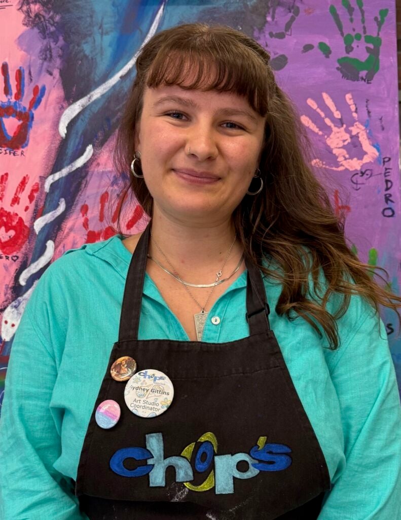 A woman wearing a black apron with "chops" logo stands in front of a colorful painted canvas with handprints.