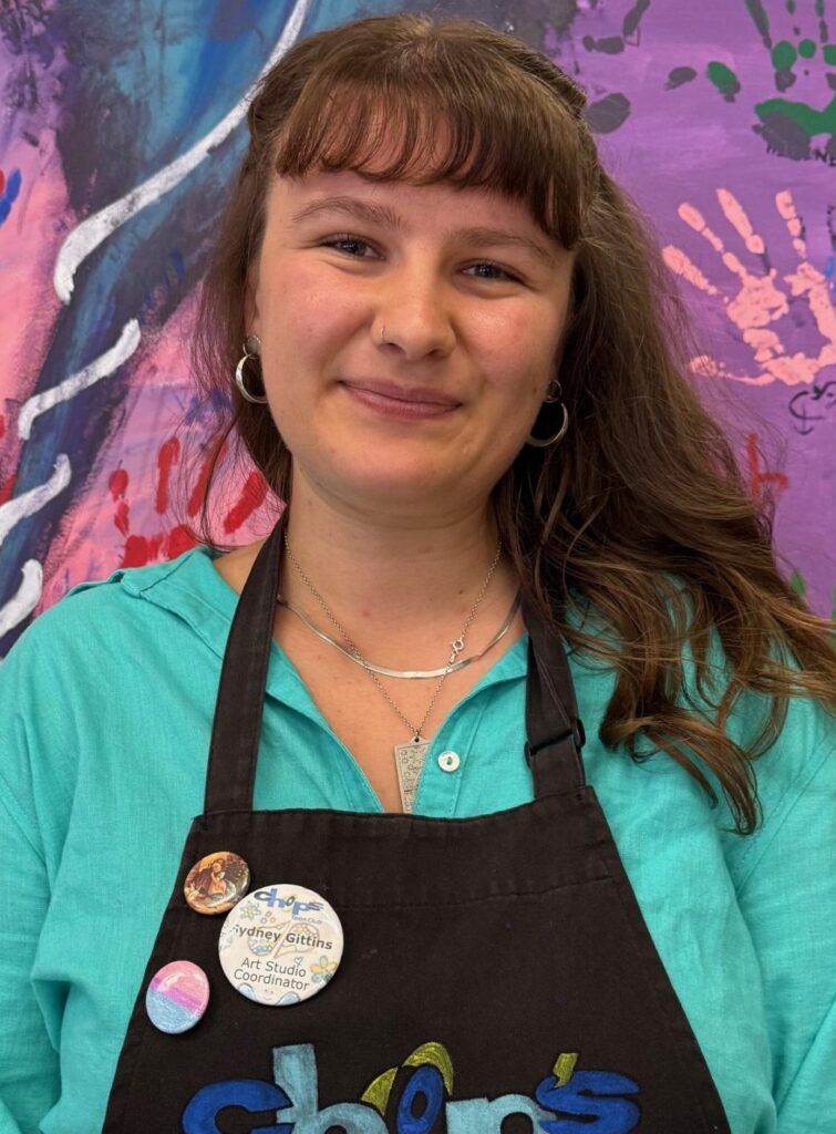A woman wearing a black apron with "chops" logo stands in front of a colorful painted canvas with handprints.